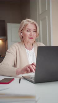 Elderly Woman Working on a Laptop and Talking on the Phone Using Wireless Headphones