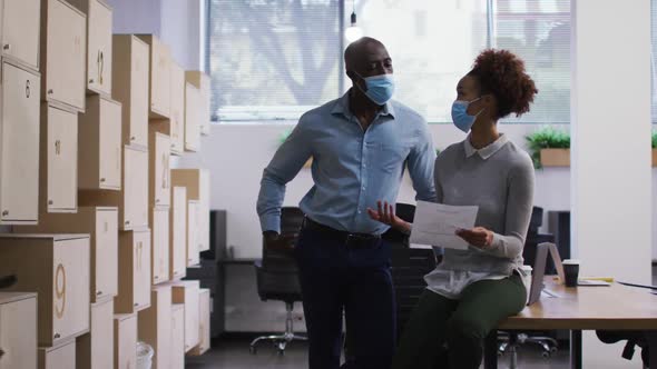 Diverse male and female business colleagues in face masks talking, woman holding document in office