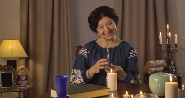 Portrait of Old Caucasian Woman in Blue Blouse Sitting at the Table with Candles and Shuffling Cards