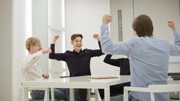 Four Joyful Millennial Male Colleagues Cheering Success Sitting at Conference Table in Office