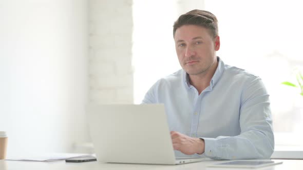 Man Looking at Camera while using Laptop in Office