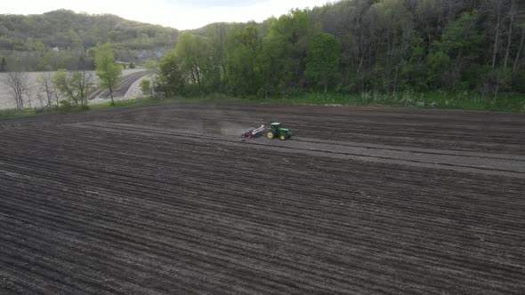 Farmer tiling field on the side of a hill with lush forest and mountains with clouds in sky.