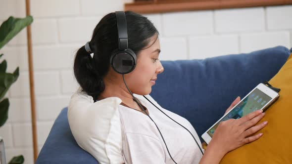 Beautiful smiling young Asian woman while on a sofa in headphones listening to music
