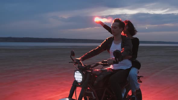 Young Couple Riding on Vintage Motorcycle with Red Burning Signal Fire After Sunset on Beach Slow