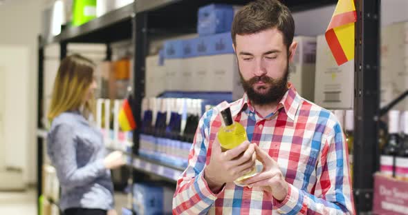 Portrait of Young Handsome Caucasian Man Examining Label on Bottle of Expensive White Wine in Shop