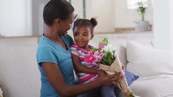 Happy african american mother and daughter sitting on sofa and smelling flowers