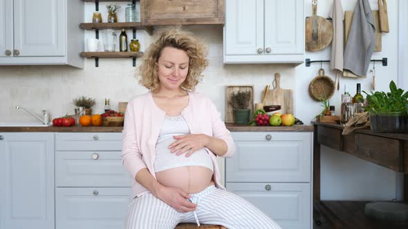 Happy Pregnant Woman Sitting On Chair And Touching Her Belly At Home