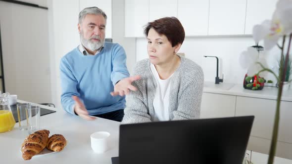 Brunette Middle Aged Woman Talking with Worried Greyhaired Senior Man Sitting in Kitchen with Laptop