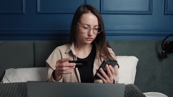 Young Woman Customer Holding Credit Card and Smartphone Sitting on Couch at Home, Happy Female