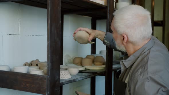 Senior Man Potter Doing Inventory Looking at Ceramics and Taking Notes in Workshop