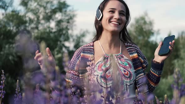 Beautiful Young Happy Woman in Headphones Dancing in a Purple Field of Lupine Flowers