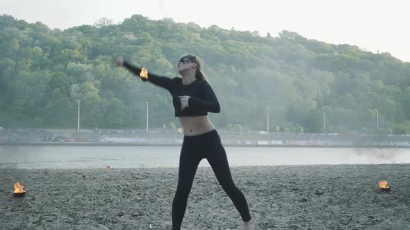 Confident Slim Woman in Mask Dancing, Performing a Show with Fireball Standing on the Riverbank