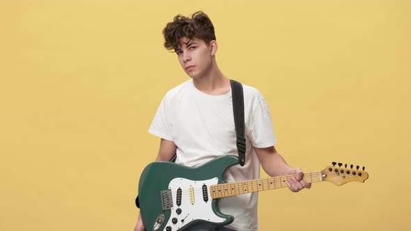 Portrait of Teenage Boy with Short Curly Hair Posing with Electric Guitar on Stage Over Yellow