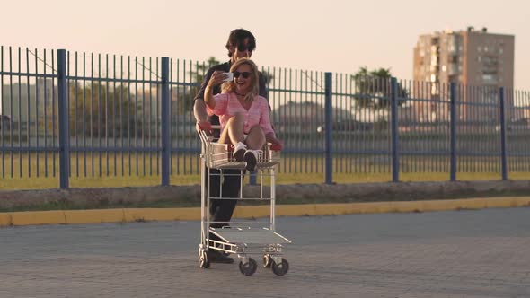 the Guy Rolls Attractive Young Woman in the Trolley on Parking Lot of the Supermarket at Sunset