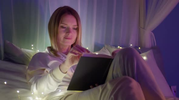 Calm Relaxed Young Woman Reading Book with Magic Lights in Cozy Home Atmosphere