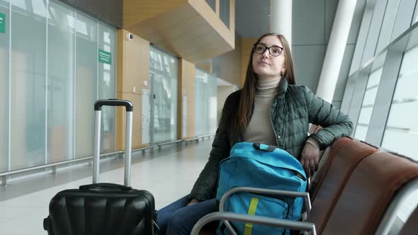 Young Tired Woman in Glasses Is Waiting Her Flight in Airport Sitting in Hall