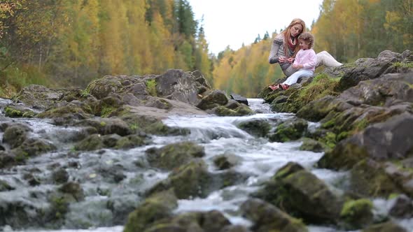 A Young Mother and Her Cute Daughter Sitting Near the Forest Stream