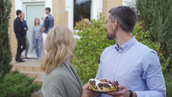 Happy Mature Couple with Cake Smiling and Looking at Camera Standing in the Foreground While Company