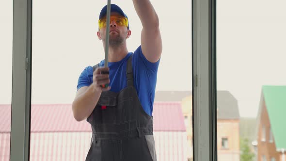 Man in Uniform Measures Size of Window in Cottage Outdoors