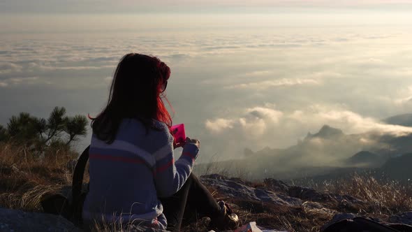 Young Redhead Woman in Sunglases Sitting on a Rock High in the Mountains Above the Clouds and