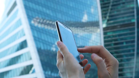 Female Hands Are Using a Smartphone Against the Background of Large Glass Business Centers, Close-up