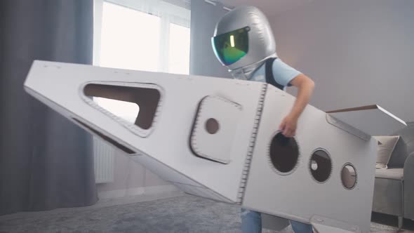 Kid Boy in an Astronaut Costume Plays with a Cardboard Space Shuttle Runs Around the Room Slow