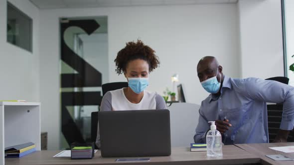 Diverse businessman and businesswoman in face masks discussing, using laptop in office