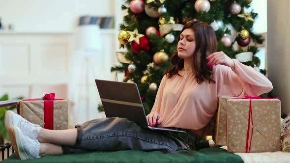 Young Woman Lying Under Decorated Pine Tree Among Gift Boxes and Packages in Home Interior