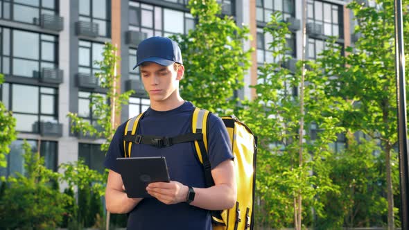 Portrait Young Man Courier Food Delivery with Thermal Backpack Uses Tablet