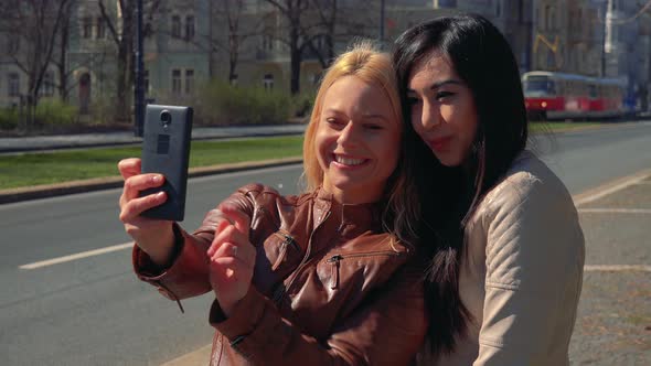 A Young Caucasian Woman and a Young Asian Woman Take a Selfie Together with a Smartphone in a Street