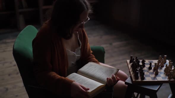 Top View of Serious Young Intelligent Woman in Glasses Reading Old Book Sitting on Chair Near Table