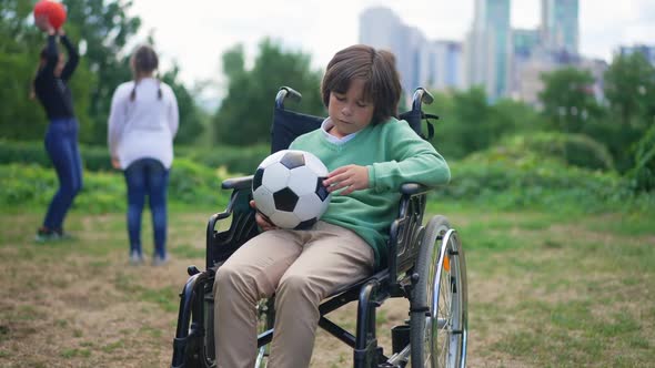 Portrait of Hopeless Caucasian Disabled Boy in Wheelchair Sitting with Ball Looking at Camera with