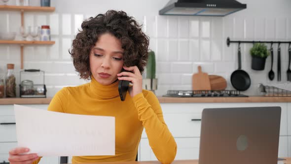 Businesswoman Talking on Mobile Phone with Financial Report in Hand in Kitchen