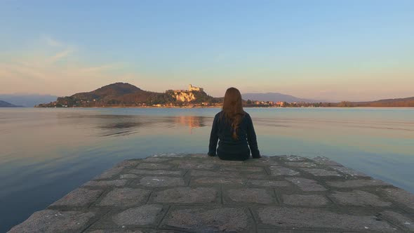 Back view of child girl sitting on pier edge looks at Angera castle on Maggiore lake in Italy at sun