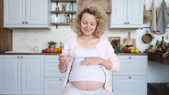 Young Pregnant Woman Holding Glass Of Water Resting In Kitchen