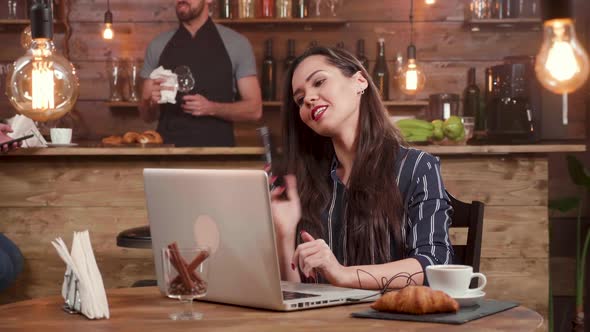 Pretty, Brunette Woman Answers a Phone Call While Working on Her Laptop Remotly