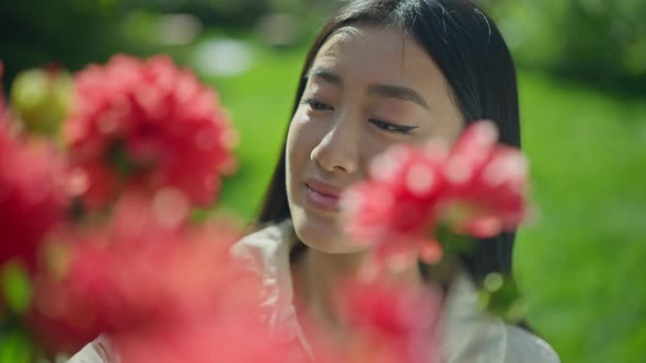 Closeup Face of Happy Young Asian Woman with Brown Eyes Behind Red Blooming Flowers in Sunny Garden