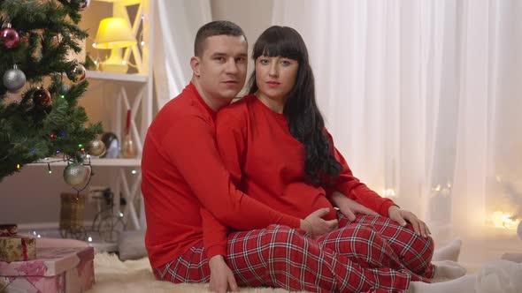 Portrait of Caucasian Man and Pregnant Woman in Similar Red Pajamas Sitting Indoors on New Year's