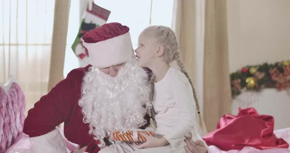 Cheerful Blond Caucasian Girl Sitting on Santa Claus Knees and Whispering on His Ear. Pretty Child