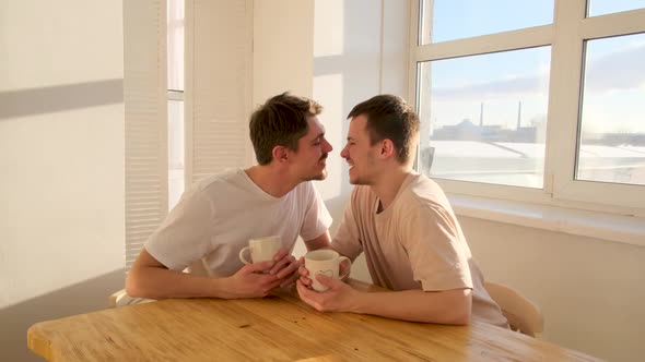 Lovely Gay Couple Kisses Sitting at Wooden Table Near Window