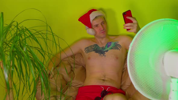 Happy Man in Santa Hat with Bare Torso Takes Selfie on Smartphone Sitting in Armchair Before Fan