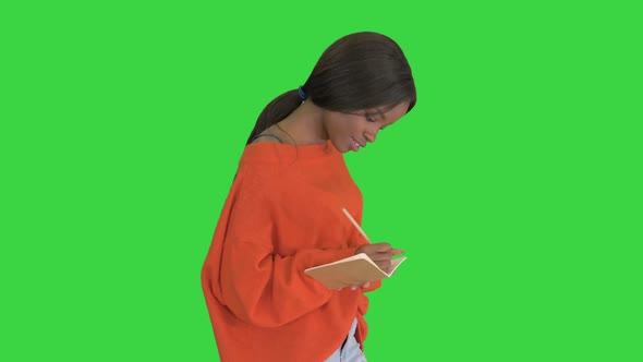 Pretty African American Woman in Bright Jumper Writing in Her Notepad While Walking on a Green