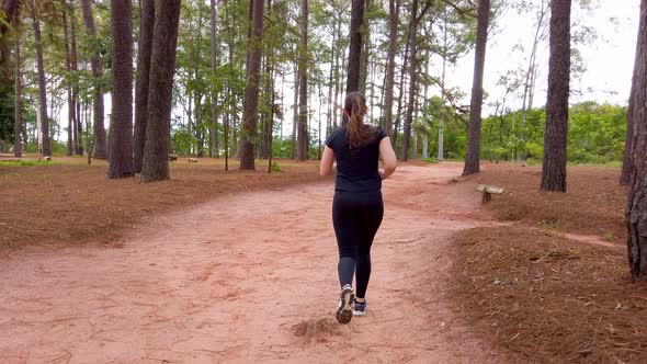 Capture following a girl jogging outdoors on a dirt trail beteen trees.