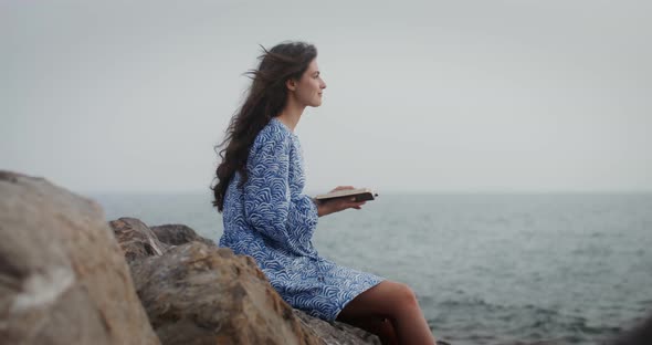 A Beautiful Young Woman is Reading a Book Sitting on a Large Stone on Seashore