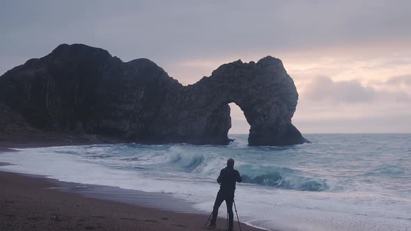 A Male Photographer With Camera On A Tripod Taking Photo Of The Natural Limestone Arch Durdle Door O