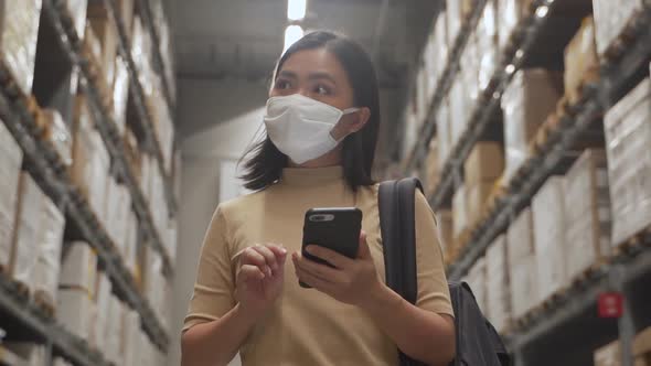 Asian woman wearing mask walking in warehouse and use smart phone.