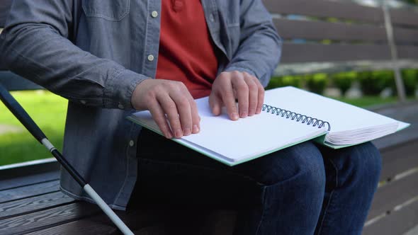 Young Blind Man Sitting on Bench in City Park and Reading a Braille Book