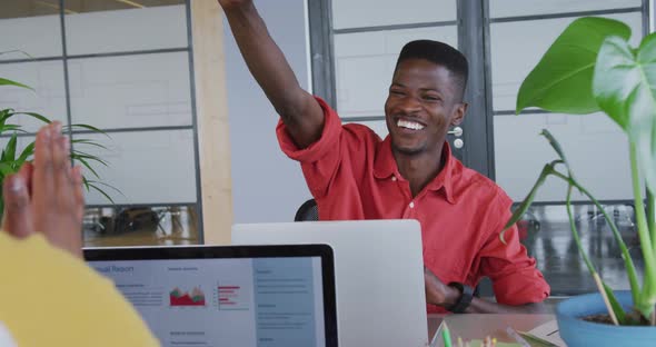 Happy african american creative businessman high fiving with female colleague at desk in office