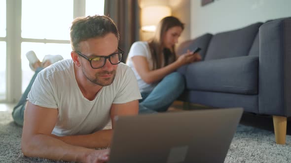 Couple Spends Time Together in a Cozy Room: Man Lies on a Carpet and Uses a Laptop, Woman Using