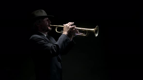 Male Musician in Hat and Classic Suit Starts Playing Musical Trumpet in Studio on Black Background